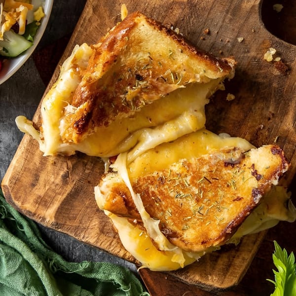 Two halves of a golden-brown grilled cheese sandwich with melted cheese and herbs on a wooden board.