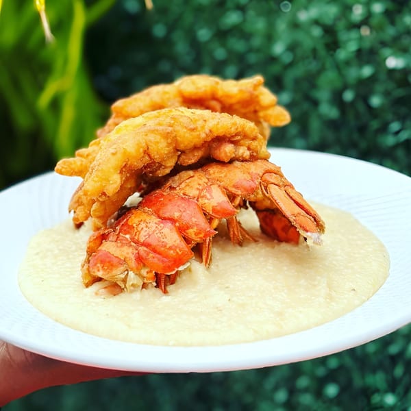 Scrum-did-dly-ump-tious's fried lobster tails and grits on a white plate with green foliage.