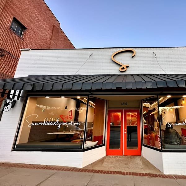 Exterior of Scrum-did-dly-ump-tious restaurant with white brick, black awning, and red double doors.