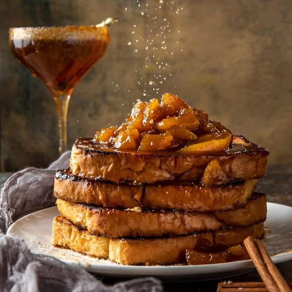Stack of Scrum-did-dly-ump-tious French toast topped with fruit and syrup, with a drink nearby.