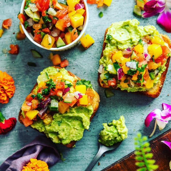 Two avocado toasts topped with colorful fruit salsa, a bowl of salsa, and flowers.